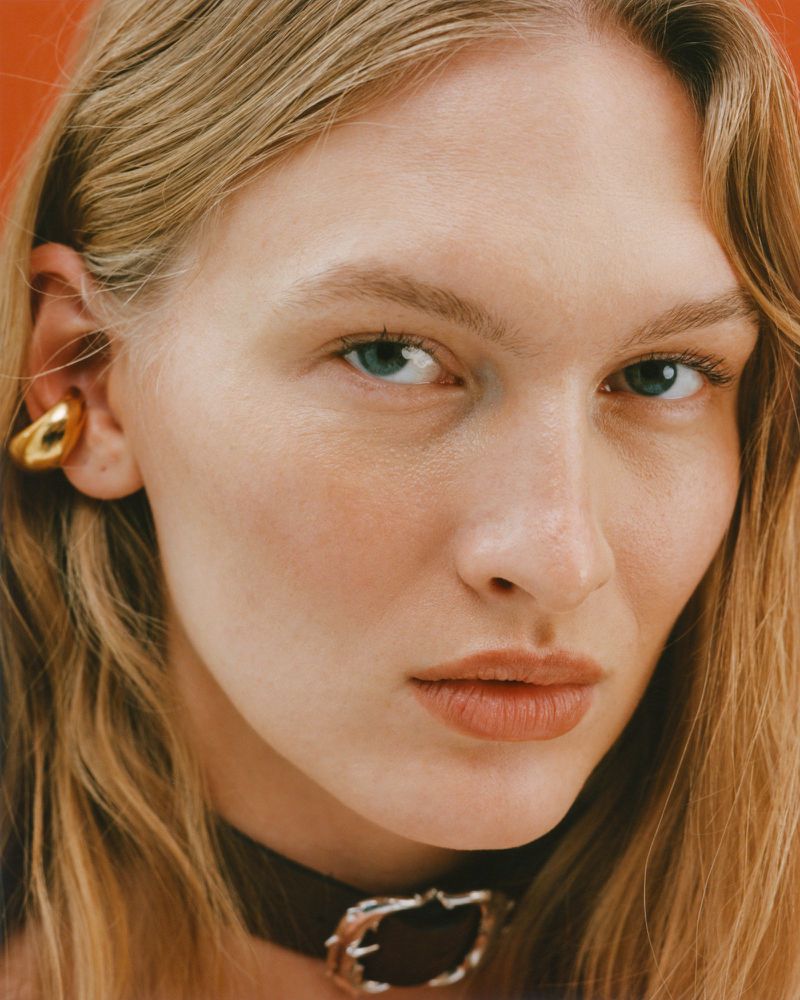 Close-up studio shot of a fair-skinned adult female model with blonde hair, wearing a dark choker and a gold ear cuff against an orange background.