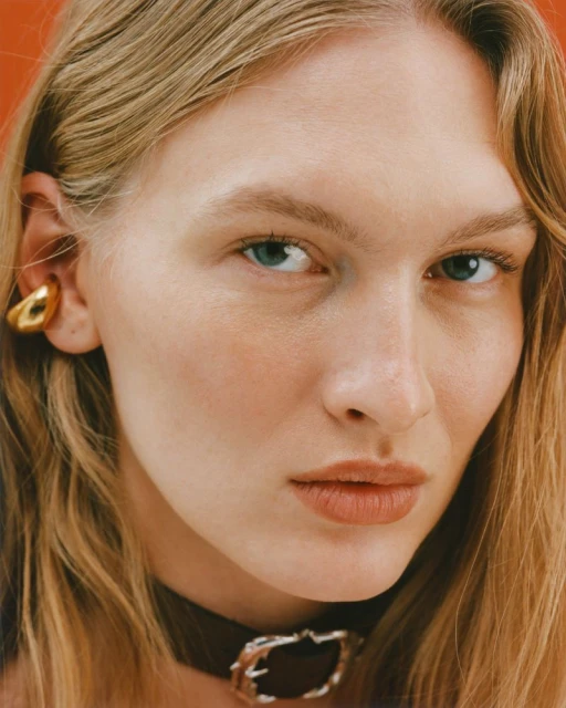 Close-up studio shot of a fair-skinned adult female model with blonde hair, wearing a dark choker and a gold ear cuff against an orange background.