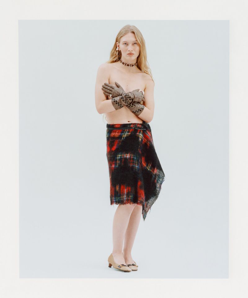 Fashionable woman in gloves and plaid skirt poses against a light backdrop in a studio, showcasing a modern editorial style.