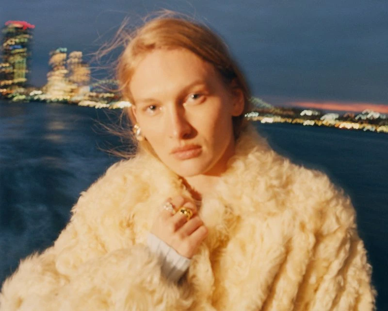Portrait of a blonde adult female wearing a cream-colored faux fur coat, with cityscape and waterfront lights blurred in the background.