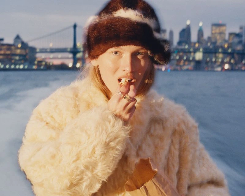 Blonde woman in fur hat and coat eating popcorn on a boat at night, bridge and city skyline in background, studio lighting and color grade.