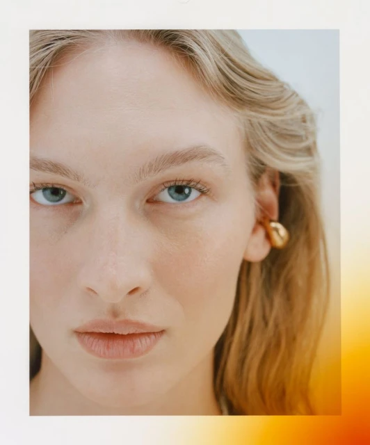 Close-up portrait of an adult woman with blonde hair and blue eyes, wearing a gold earring, in a beauty shot with neutral background.