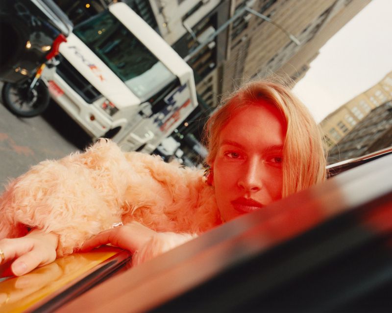 A woman in a fur coat leans out of a car window in a tilted shot in an urban environment, with a truck and buildings in the background.