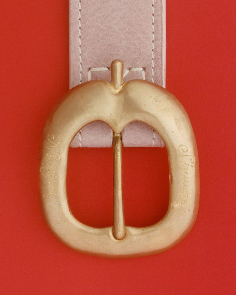 Close-up of a light pink leather belt with a gold apple-shaped buckle on a red background.