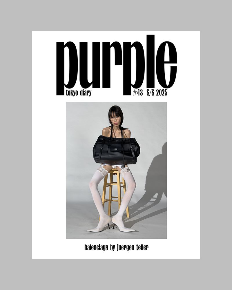Magazine cover featuring a slender adult female in white stockings, balancing a large black bag while seated on a wooden stool.