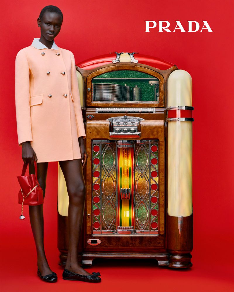 Fashion editorial shot of a Black female model in a peach coat, red purse, and black flats standing next to an ornate jukebox with a red background.