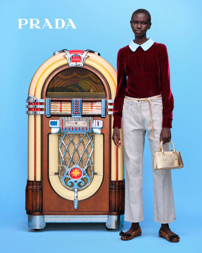 A Black woman poses in a fashion shot, wearing a burgundy top, white jeans, and animal-print shoes, standing next to a vintage jukebox.