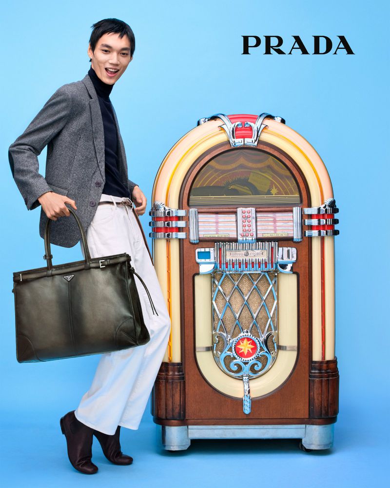 A slender male model wearing a grey jacket and white trousers is holding a black leather Prada handbag next to a retro jukebox on blue.
