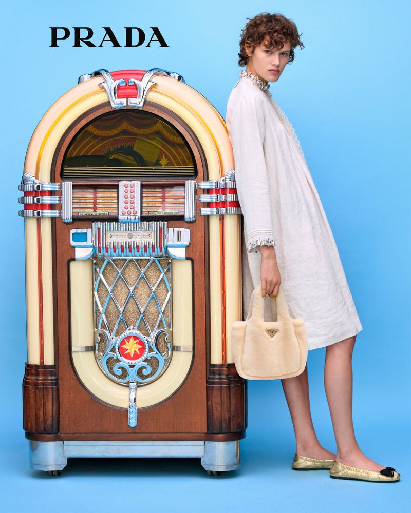 Fashion shot featuring a model in a white dress with a beige handbag next to a vintage jukebox against a light blue background.