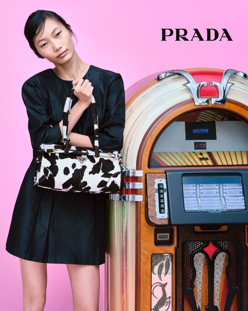 A woman poses with a Prada purse next to a jukebox in a studio shot against a pink background, highlighting fashion and vintage charm.