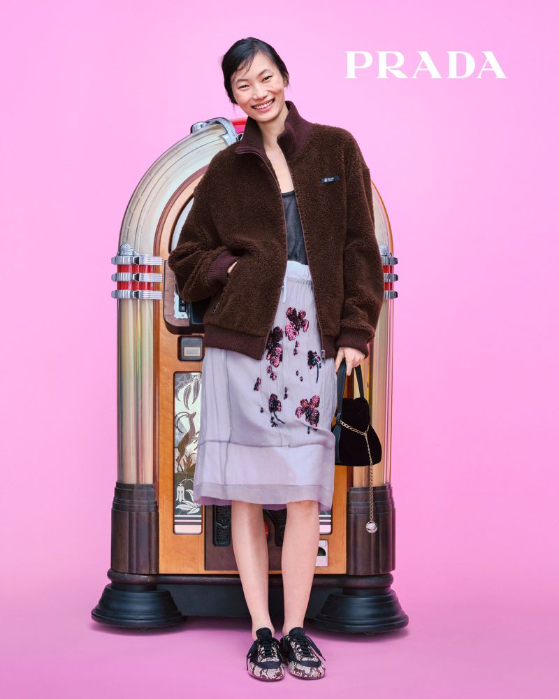 An Asian woman in a brown jacket, light-colored skirt, and black shoes stands next to a vintage jukebox against a pink background, promoting Prada.