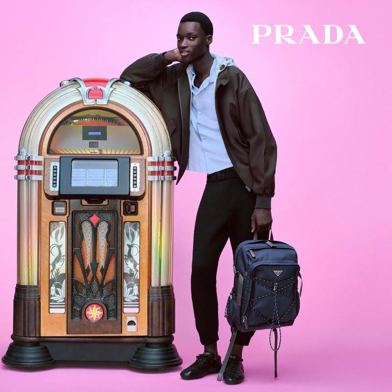 Stylish male model poses with vintage jukebox against pink backdrop while holding a blue backpack in a fashion advertisement.