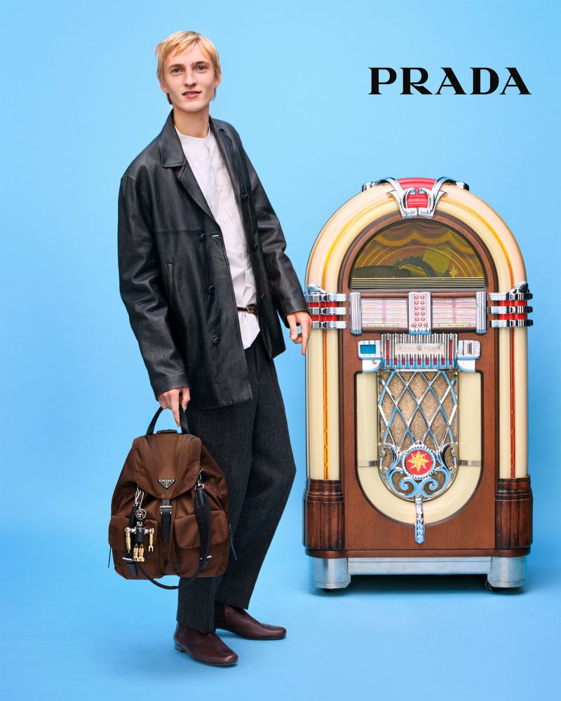 A stylish man in a jacket stands next to a vintage jukebox, holding a brown backpack with the Prada logo, set against a blue background.