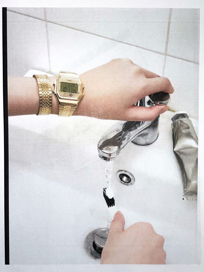 Close-up of a person washing a toothbrush with water from a faucet in a white sink, wearing a gold watch and bracelet.