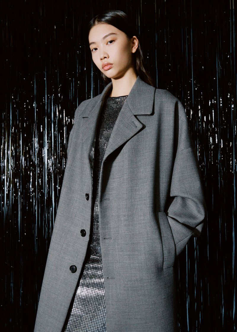 Asian woman poses in grey wool coat and glittering mini dress against a black background with shiny silver streamers, fashion shoot.