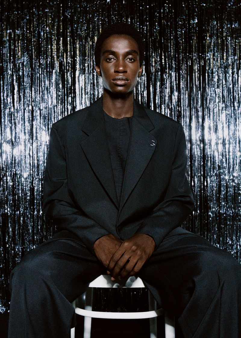 A dark-skinned adult male model in a black suit sits on a white stool in front of a silver, sparkling background, posing for a studio portrait.