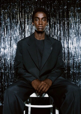A dark-skinned adult male model in a black suit sits on a white stool in front of a silver, sparkling background, posing for a studio portrait.