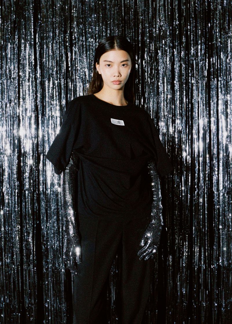 Adult woman posing in a black t-shirt and pants with long, shiny black gloves against a shiny, silver metallic fringe backdrop.