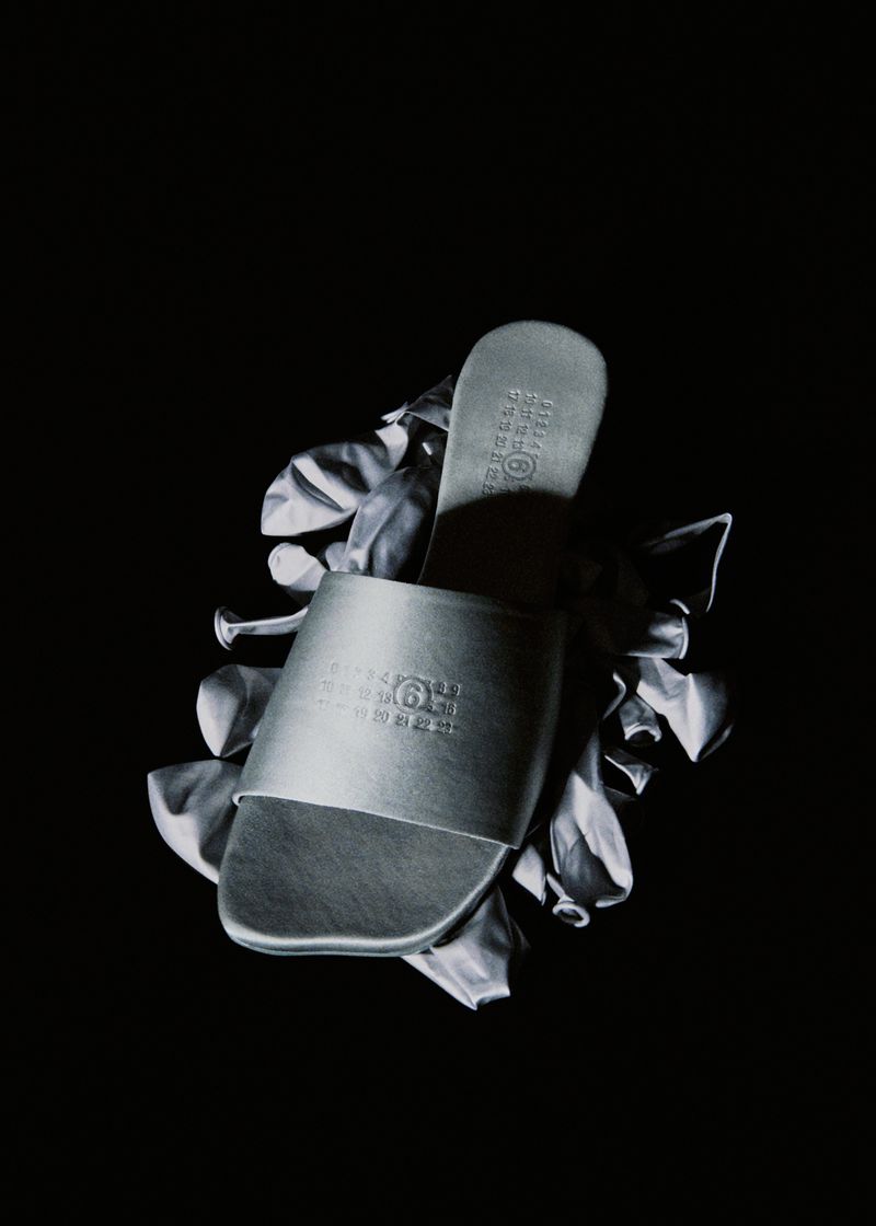 Fashion shot of a silver slide sandal featuring brand logo, resting on deflated silvery balloons against a stark black backdrop.