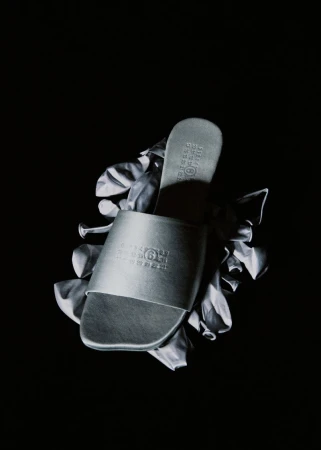 Fashion shot of a silver slide sandal featuring brand logo, resting on deflated silvery balloons against a stark black backdrop.