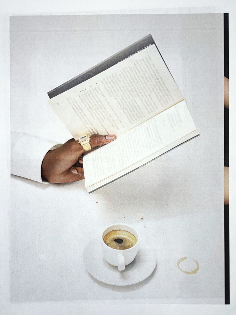 A hand holds a book above a table with a cup of coffee and a coffee stain ring, against a clean white background.