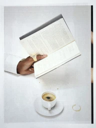 Overhead view of a hand wearing a gold watch ring holding an open book above a cup of espresso on a light surface.