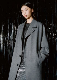 East Asian woman wearing an oversized grey tailored coat over a sparkling metallic dress against a shimmering black tinsel backdrop.
