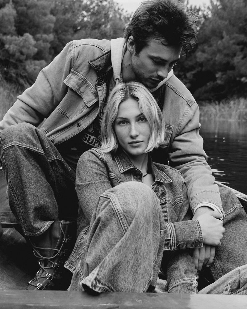 Black and white photograph of a couple in a boat on a lake, surrounded by trees. Both are wearing denim jackets and jeans.