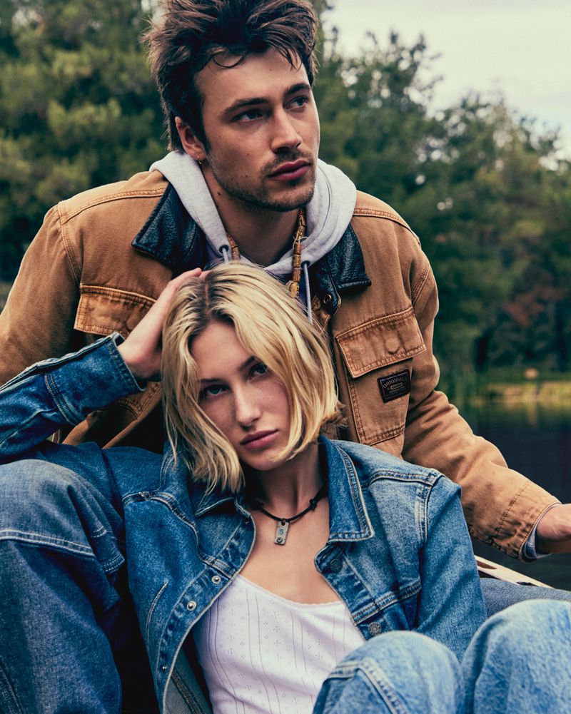 Man in brown jacket and woman in denim jacket posing outdoors together for a Guess campaign
