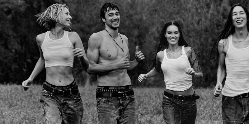Four models in white tank tops and denim jeans running outdoors in a field, captured in a black and white campaign image.