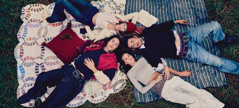 Four friends lie on patterned blankets in a grassy outdoor setting, wearing casual clothes, in a eye-level medium shot.