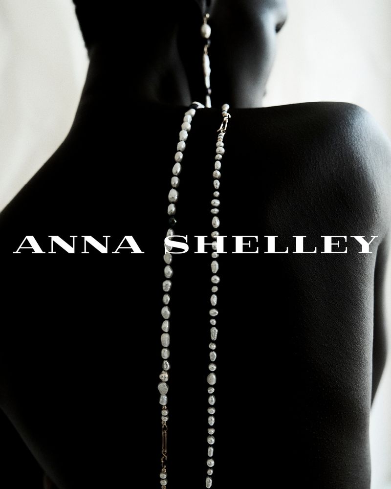 Elegant back view of a woman with dark skin wearing long pearl earrings and a necklace against a soft white backdrop in a beauty shot.