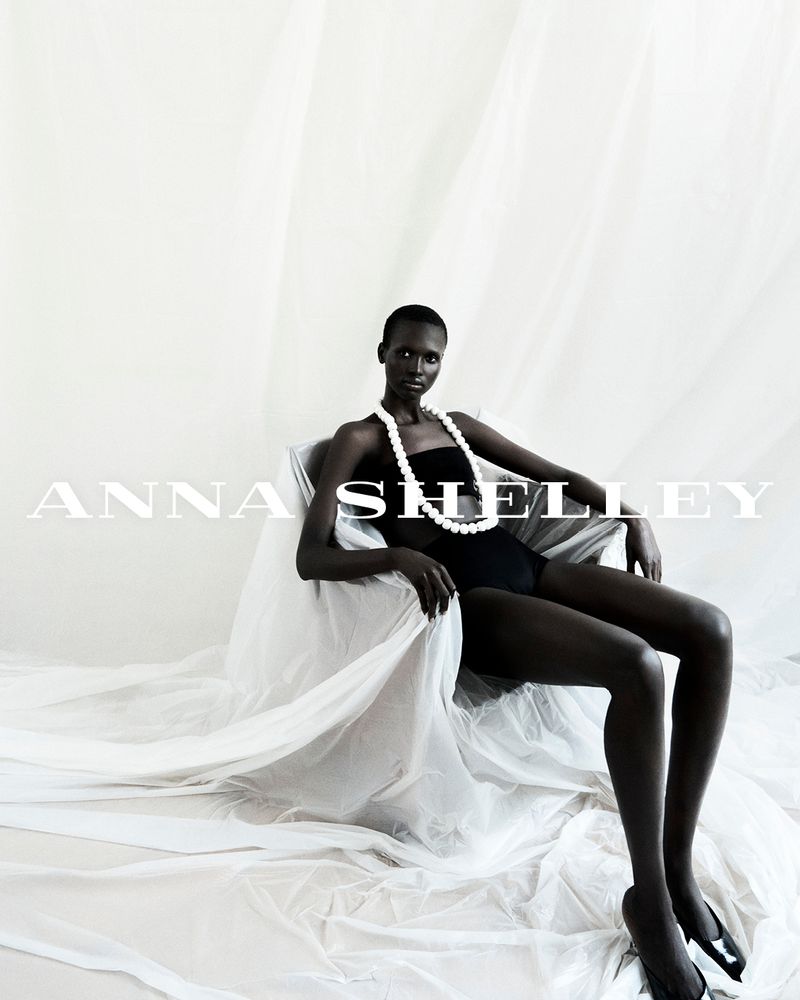 Elegant dark-skinned adult female posing in a chair with drapery, wearing a black strapless dress and pearl necklace in a studio.
