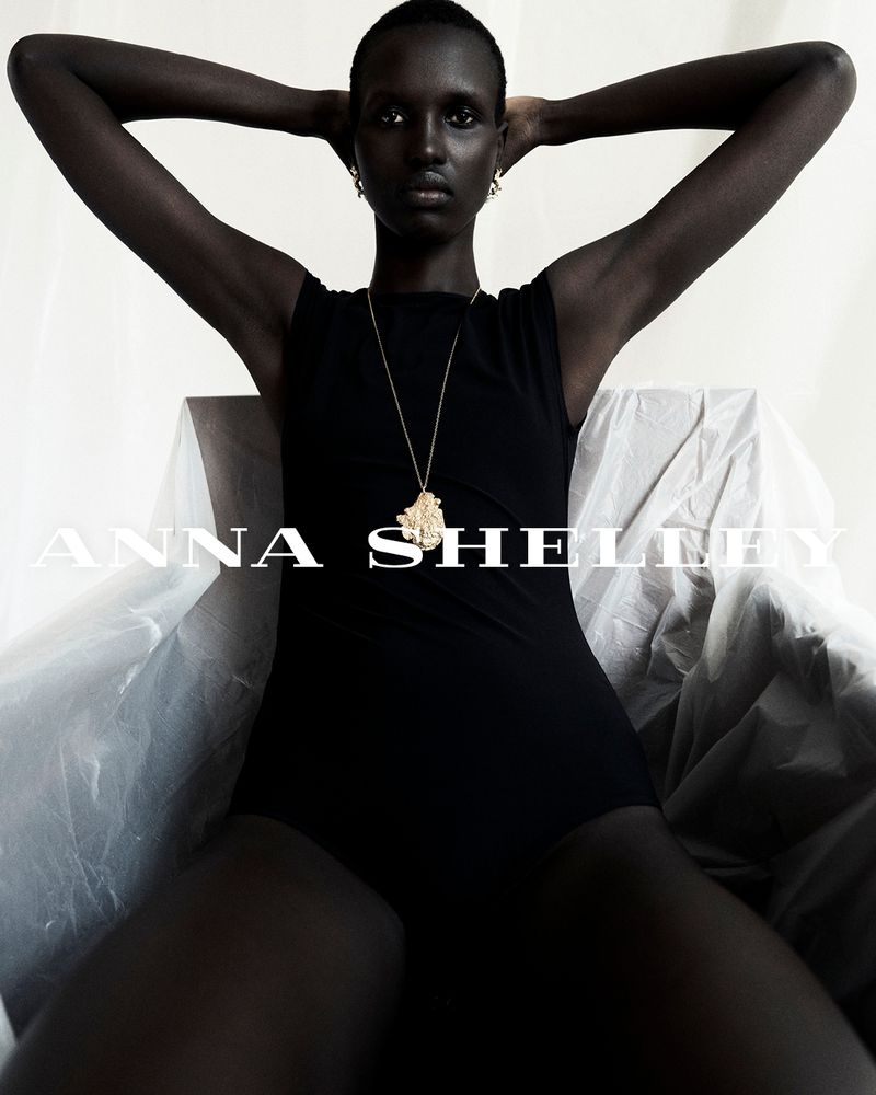 Portrait of a striking dark-skinned fashion model in a black bodysuit, posed against a light backdrop, accentuated with a gold pendant necklace.