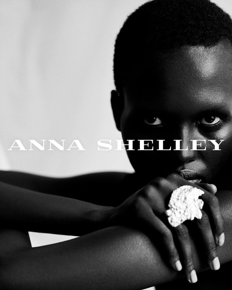 Close-up black and white portrait of an adult female with dark skin wearing a statement ring, hands folded beneath her chin.