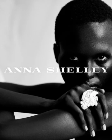 Close-up black and white portrait of an adult female with dark skin wearing a statement ring, hands folded beneath her chin.
