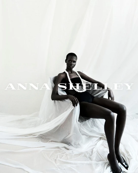Model Amar Akway seated in a draped white chair wearing black attire and a pearl necklace for Anna Shelley Fragment Collection 2.