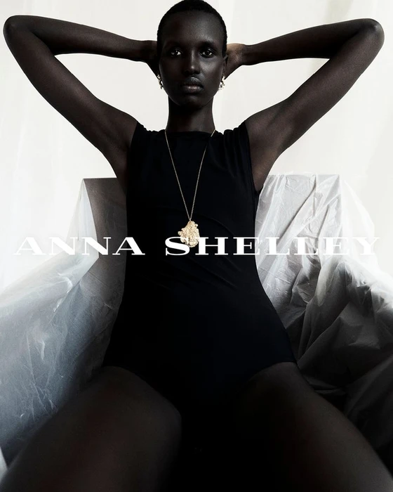 Model Amar Akway wearing a black top and gold pendant necklace seated against a draped white background