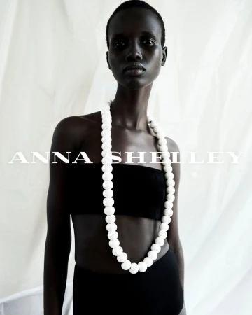 Amar Akway wearing a long white beaded necklace and black bandeau top against a draped white backdrop.