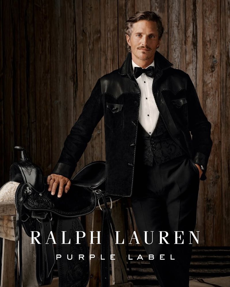 Elegant adult male in black suit and bow tie poses with a dark-colored western saddle against a rustic wooden backdrop.