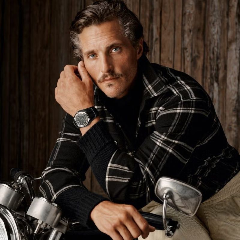 Portrait of a mature adult male with a mustache, wearing a black-and-white plaid shirt and a watch, posing with a motorcycle.