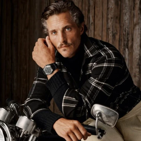 Portrait of a mature adult male with a mustache, wearing a black-and-white plaid shirt and a watch, posing with a motorcycle.