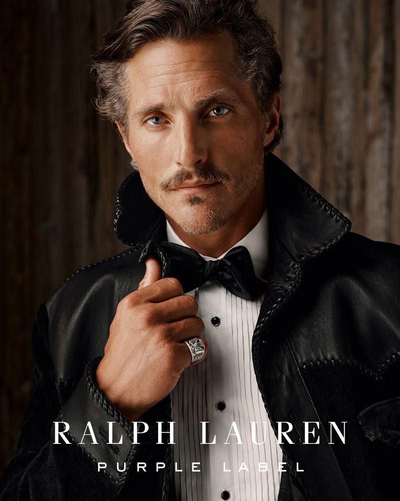 Man in tuxedo with bow tie and black jacket. Ralph Lauren Purple Label advertisement against a wood panel background.