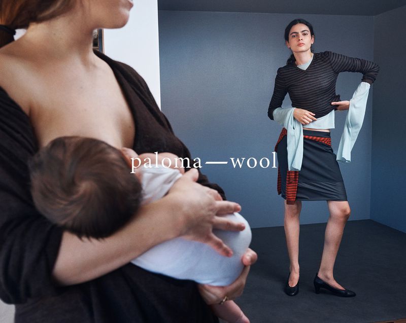 Mother breastfeeding baby indoors next to fashion model in striped top and skirt, posed in front of a blue-grey wall.