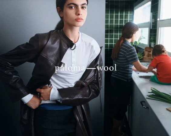 A woman models a dark-brown leather coat with a white shirt. Two figures can be seen in the background, inside a kitchen setting.