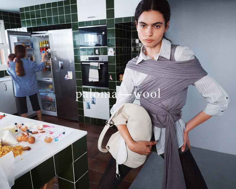 Indoor fashion shot features a young woman in a wrapped grey shawl holding a handbag in a kitchen, with a woman at the refrigerator.