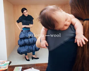 Model wearing a blue puff skirt and dark top in a wooden room, viewed past a person holding a baby