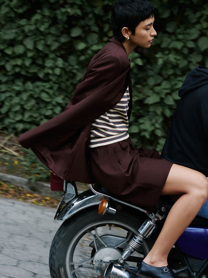 A woman with short dark hair rides a motorcycle in a brown suit with a passenger in an outdoor setting with greenery behind them.