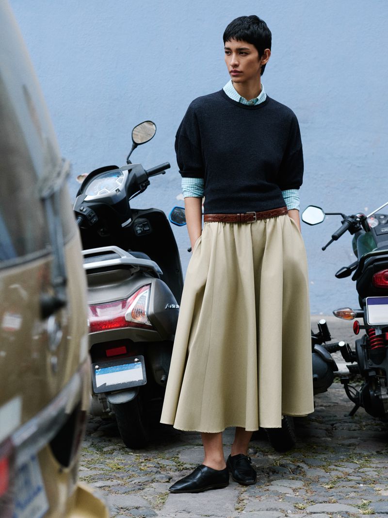 Fashion shot of a woman in a long skirt, black sweater, and black shoes, standing next to motor scooters on a cobblestone street.