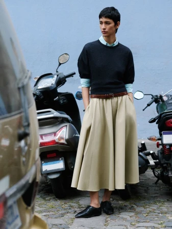 Fashion shot of a woman in a long skirt, black sweater, and black shoes, standing next to motor scooters on a cobblestone street.